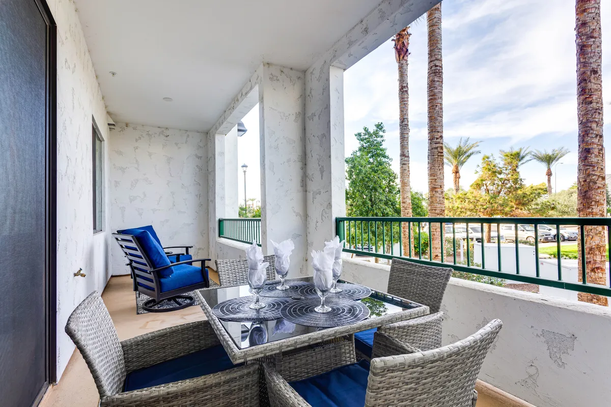This image depicts an inviting outdoor patio space featuring a dining table with blue chairs set against a backdrop of palm trees and a decorative railing. Lush greenery surrounds the area, creating a tranquil and private atmosphere ideal for outdoor dining, morning coffee, or relaxing with a book.
