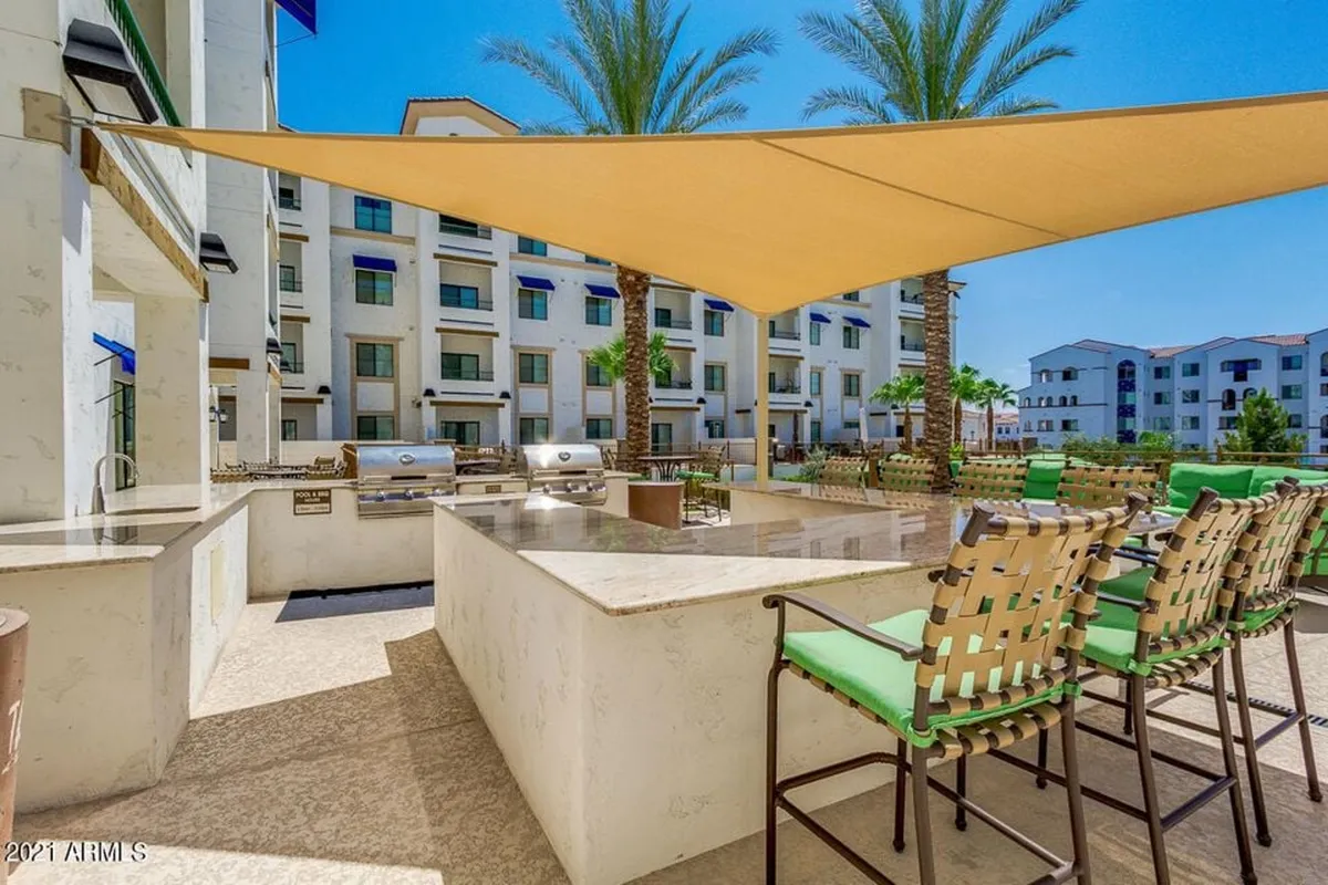 A bright exterior scene featuring a grill area with a modern seating arrangement under a shade sail, surrounded by palm trees. The setup encourages outdoor dining, socializing, and community barbecues.