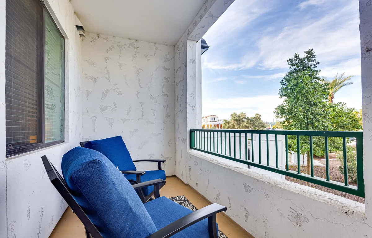 A serene balcony area with blue chairs and a green railing overlooking lush landscaping. The seating arrangement invites relaxation and quiet moments while taking in views of the surrounding greenery.