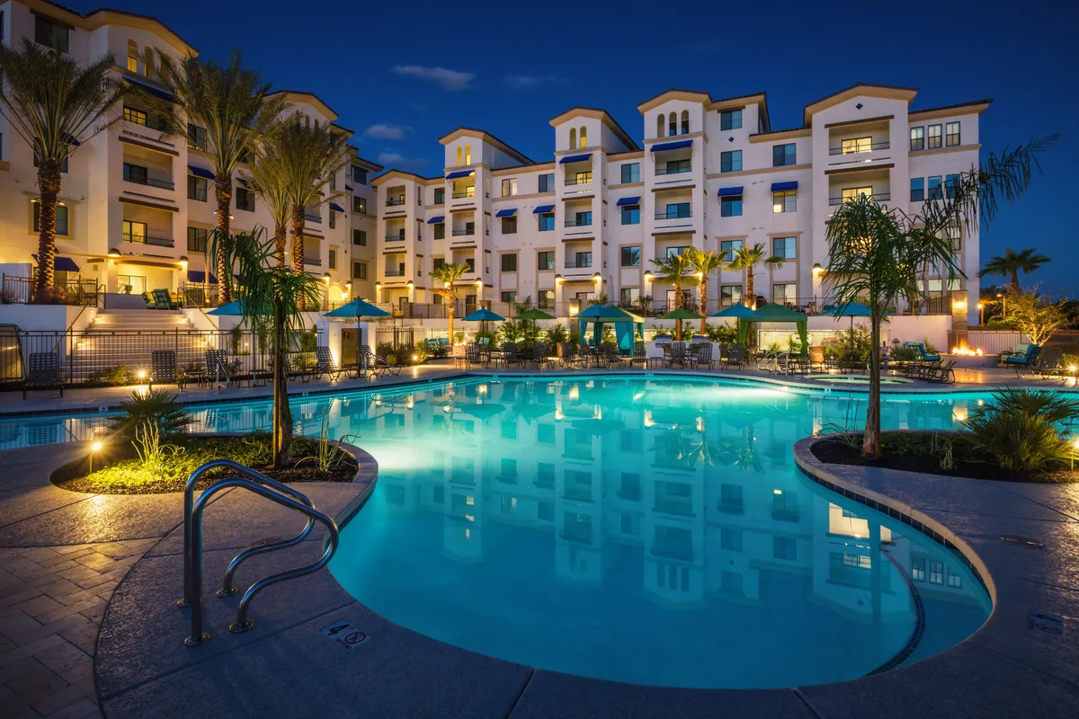 A luxurious resort-style swimming pool surrounded by palm trees and illuminated building facades. The scene includes lounge chairs and canopy-covered relaxation areas, setting a tranquil and upscale atmosphere for residents and guests. Subtle evening lighting enhances the sense of resort living and social gathering potential.