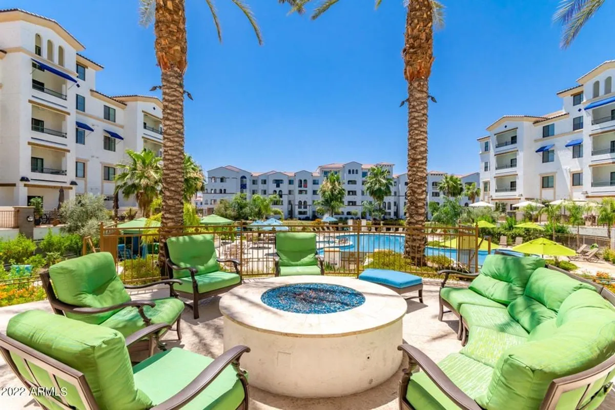 An inviting exterior scene showcasing a stylish fire pit surrounded by green chairs, a sparkling swimming pool, tall palm trees, and the backdrop of modern buildings. The area is designed for both relaxation and social gatherings, creating a lively yet comfortable atmosphere.