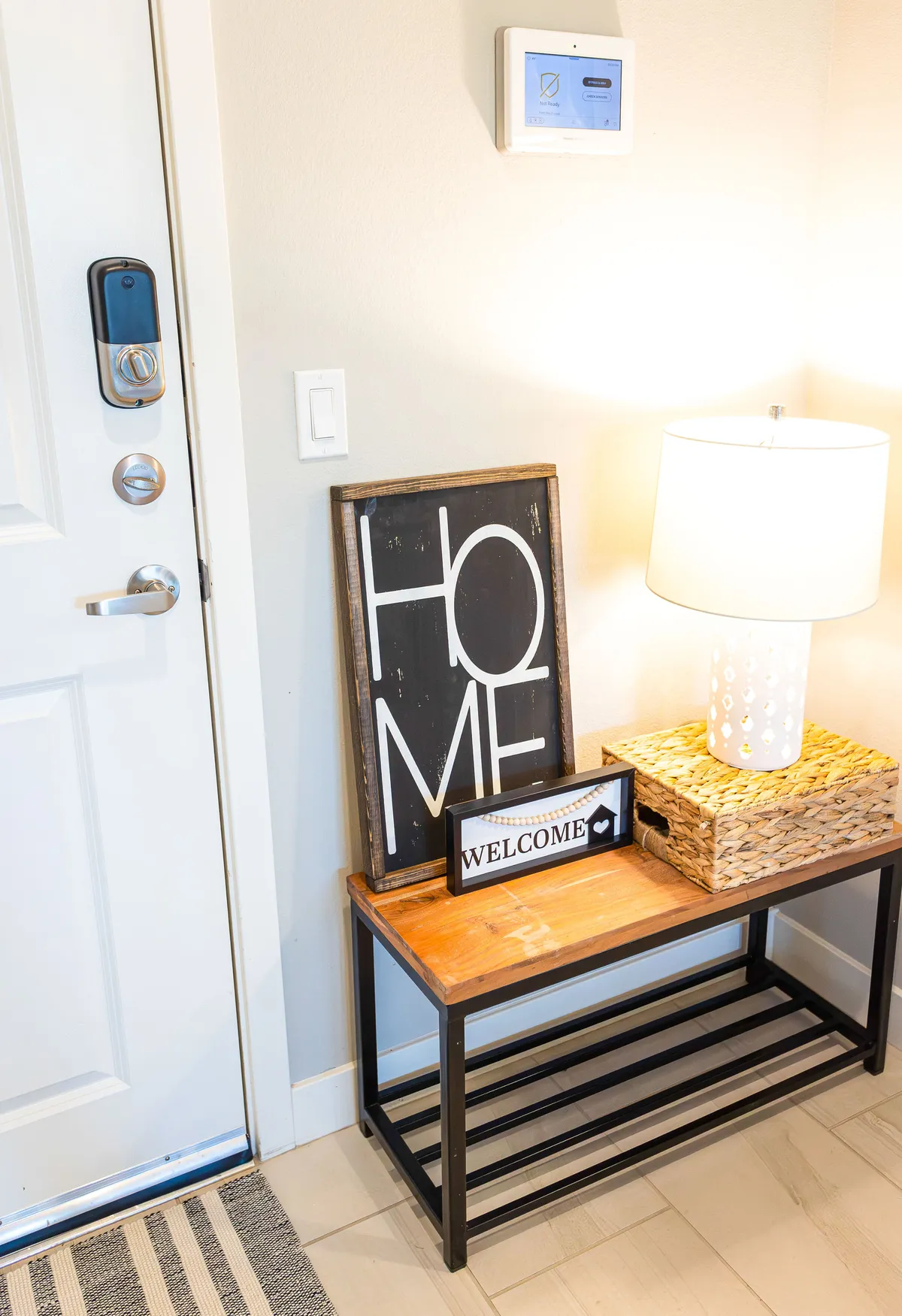 The image shows a welcoming entryway with a modern door lock, a 'HOME' sign, and a 'WELCOME' sign, next to a lamp and a woven storage basket. The scene feels inviting and homey, evoking the comfort and belonging of a long-term residence.