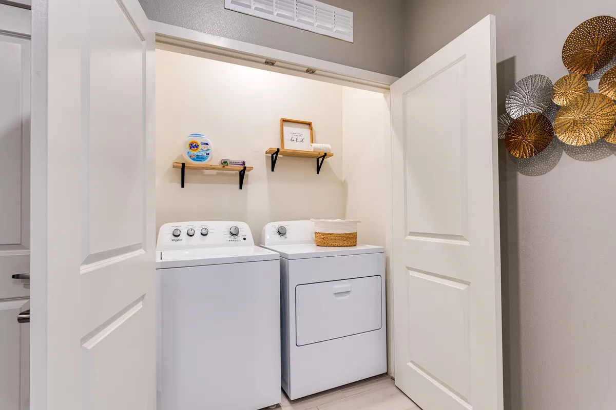 A practical, well-organized laundry area featuring a washing machine, dryer, shelves, and tasteful decorative wall art. The space is clean and accessible, emphasizing convenience for daily living.