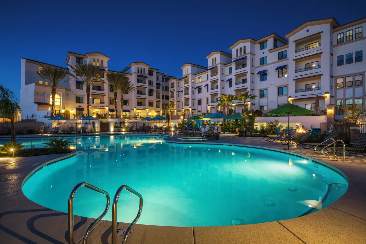 An illuminated swimming pool at night, surrounded by palm trees and outdoor seating areas. The balconies of the adjacent building overlook the pool, and the scene glows with ambient lighting, suggesting luxury and sociability after sundown.