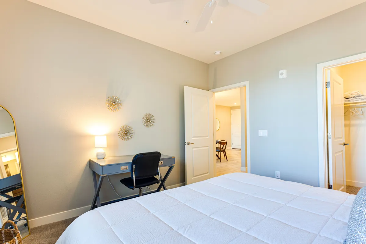 A calm and productive space featuring a bed with a white quilt, a functional desk with a chair positioned beside it, and tasteful wall accents. The setting is peaceful and well-suited for both rest and remote work.