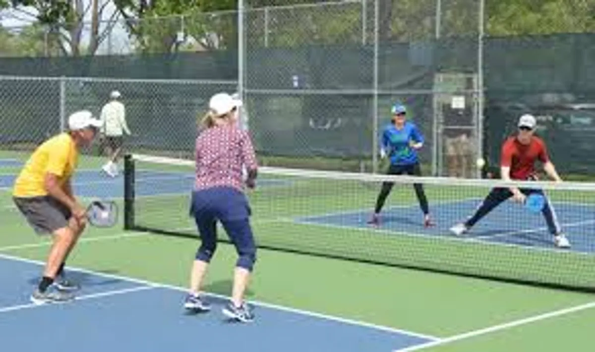 An exterior shot of a tennis court, with players actively engaged in a game. The court is surrounded by a well-maintained environment, and the scene exudes a sense of community, activity, and outdoor enjoyment. People are socializing and practicing their tennis skills in the pleasant weather.