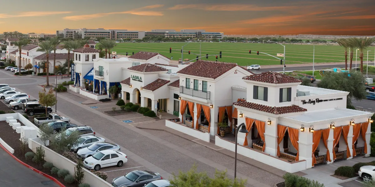 An exterior view of vibrant restaurant facades adorned with orange drapery, palm trees, and a parking area. The scene buzzes with the energy of outdoor dining and community activity, evoking a lively, welcoming atmosphere.