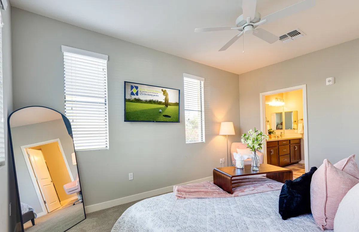 A spacious bedroom with a large bed topped with decorative pillows, a flat-screen TV, a mirror, a window with blinds allowing in natural light, and a bedside lamp. The room balances comfort and modern touches.