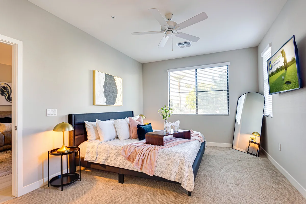 A tranquil bedroom with a comfortable bed adorned with decorative pillows, a stylish lamp on the bedside table, a large mirror, and a television mounted on the wall. The soft lighting and serene décor create an inviting, restful atmosphere.